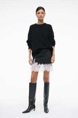 Woman wearing a black mini skirt with white lace hem, paired with a black sweater and black knee-high boots.