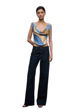 A model stands against a plain white background wearing a sleeveless draped top with blue, beige, and brown panels, paired with dark wide-leg jeans and black sandals.