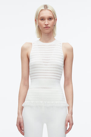 Woman with arms by her side wears white ribbed knit tank top with subtle variegated stripes and ruffled hemline.