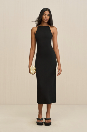 Woman wearing a black sleeveless midi dress that features a high neck, paired with black sandals and a cream clutch.