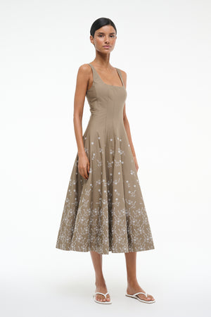 Woman wearing a taupe sleeveless midi dress that features a square neck, fitted bodice, and A-line skirt with white floral embroidery, paired with white sandals.