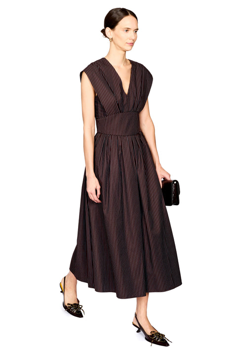 A woman modeling a sleeveless dark brown midi dress with thin vertical pinstripes, a V-neckline, and a gathered waist, paired with black pointed shoes and a small black clutch, against a plain white background.