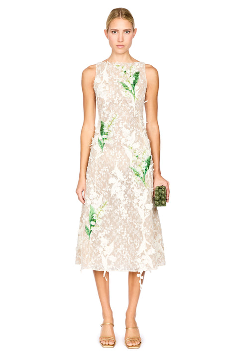 A model wearing a sleeveless cream lace midi dress with green floral applique, paired with gold high-heeled sandals, against a plain white background.
