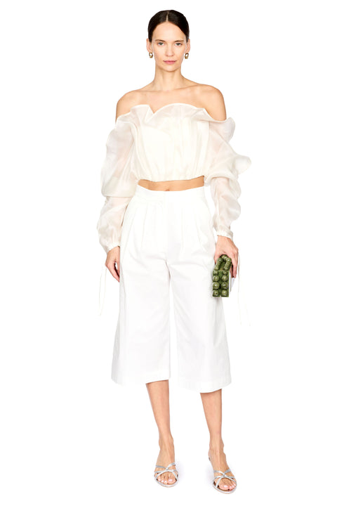 A model wearing a white off-the-shoulder crop top with dramatic puffed sleeves, matching white wide-leg cropped pants, and silver strap sandals, holding a small green textured clutch against a plain white background.