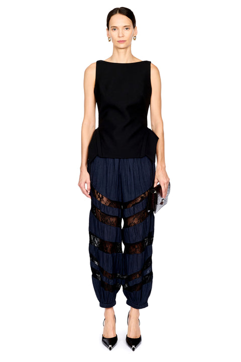 A model wearing a sleeveless black top and dark navy pleated harem-style pants with sheer black lace bands, standing against a plain white background.