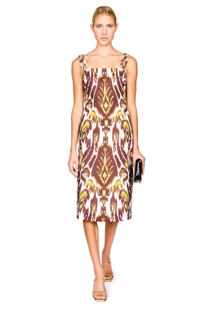 A woman modeling a midi dress with a brown, gold, and cream ikat pattern, paired with gold sandals and holding a small black clutch, set against a plain white background.