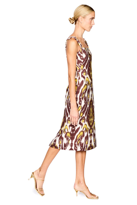 A woman modeling a midi dress with a brown, gold, and cream ikat pattern, paired with gold sandals and holding a small black clutch, set against a plain white background.