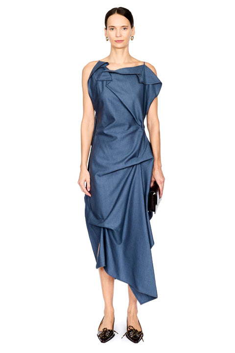 A model wearing a slate blue one-shoulder draped midi dress with a high-low asymmetrical hem, paired with black pointed flats and holding a small black clutch, set against a plain white background.