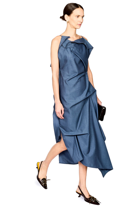 A woman modeling a draped midi dress in a dusty slate blue color, paired with black pointed-toe kitten heels and holding a small black clutch, set against a plain white background.