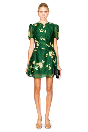 A model wearing a short-sleeved green dress with a yellow floral print, holding a small black clutch, set against a plain white background.