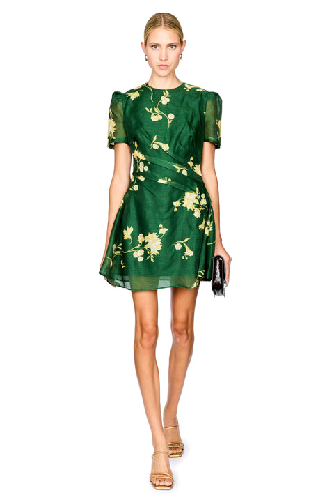 A model wearing a short-sleeved green dress with a yellow floral print, holding a small black clutch, set against a plain white background.