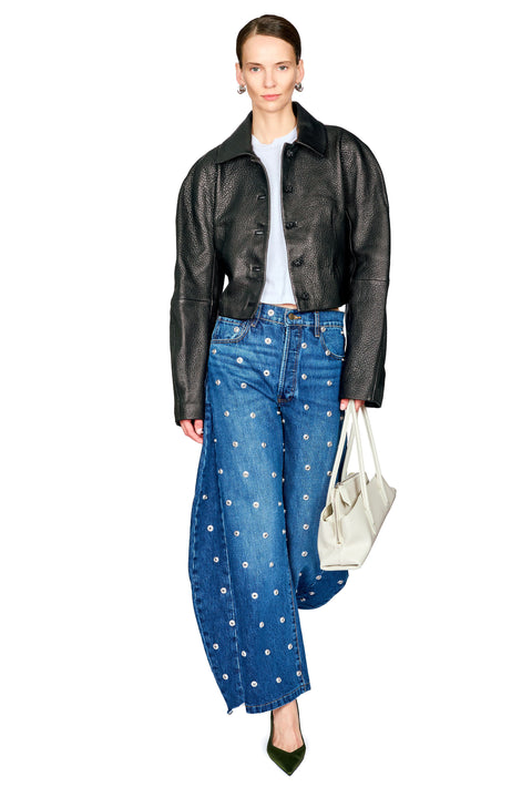 A model is shown from the head to toe wearing a black leather jacket, blue wide-leg jeans with white floral embroidery, a white t-shirt, and holding a white tote bag against a solid white background.