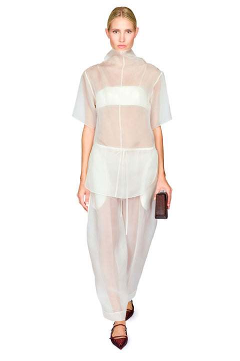 A model wearing a sheer white high-neck short-sleeve top and matching wide-leg sheer trousers, holding a small dark clutch against a plain white background.