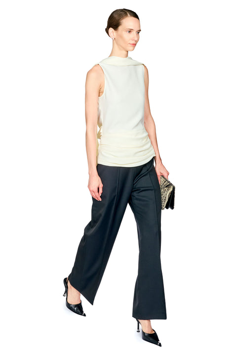 A model is shown from a side-back angle wearing a sleeveless cream mock-neck top and black wide-leg trousers, holding a small patterned clutch against a plain white background.