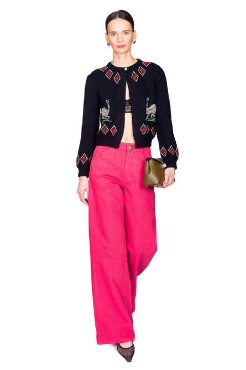 Woman wearing a black embroidered cardigan and pink pants on a white background one leg in front.