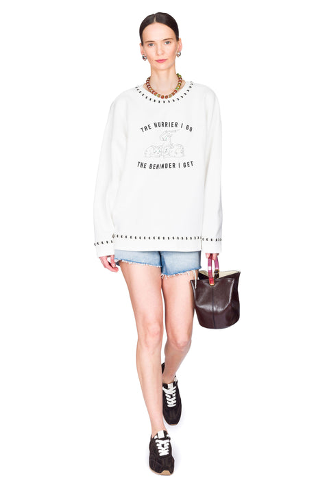 Woman wearing a white sweater with text, denim shorts, and holding a brown bag on a white background with one foot forward.