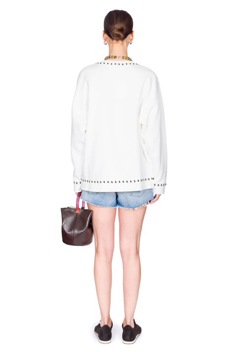 Woman wearing a white sweater with text, denim shorts, and holding a brown bag on a white background from the back.
