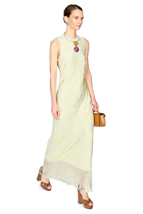 A woman modeling a sleeveless, light yellow-green maxi dress with a fringed hem, styled with a chunky statement necklace, brown platform sandals, and a small brown handbag against a plain white background.