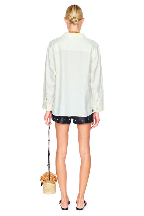 A model shown from the back wearing a cream-colored long-sleeve button-up shirt, dark shorts with white polka dots, and holding a small woven straw bag with a black strap against a solid white background.