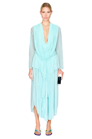 A model wearing a light aqua blue pleated long-sleeve top and maxi skirt set with a draped V-neckline, paired with colorful flat sandals and holding a black clutch, set against a plain white background.