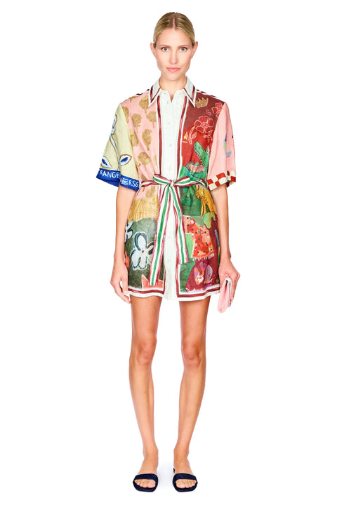 A model wearing a short-sleeved, collared mini dress with a multicolor abstract print, featuring a tie waist belt and striped trim, paired with black slide sandals against a plain white background.