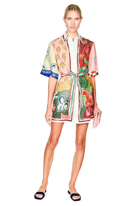 A model wearing a short-sleeved, belted shirt dress with a vibrant multicolor floral and botanical print, styled with dark blue slide sandals and holding a pink clutch against a plain white background.