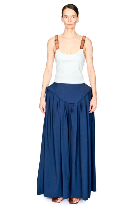 A model wearing a white ribbed tank top with brown shoulder straps and a long navy blue pleated skirt with a curved waistband, standing against a solid white background.