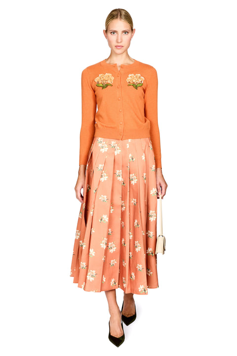 A model stands against a white background wearing a two-piece outfit consisting of a burnt orange long-sleeve button-up top with floral embroidery on the chest and a matching peach-orange pleated midi skirt with a white and green floral print. She is holding a small beige handbag and wearing black pointed-toe heels.