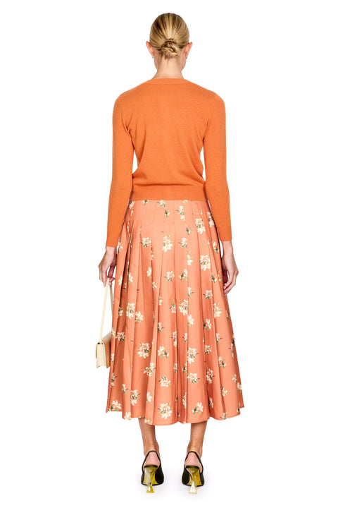 A woman shown from the back wearing a bright orange long-sleeve knit top and a peach-orange pleated midi skirt with a small white floral print. She is holding a small beige handbag and wearing black and gold high heels against a plain white background.
