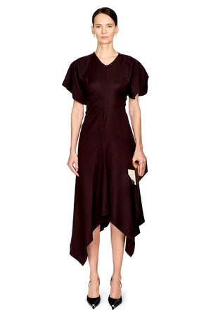 A woman modeling a dark burgundy midi dress with short puff sleeves, a V-neckline, and an asymmetrical draped hem, paired with black pointed heels against a plain white background.