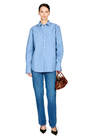 A model stands against a white background wearing a light blue long-sleeve button-up shirt, blue jeans, and black pointed heels, while holding a brown leather handbag.