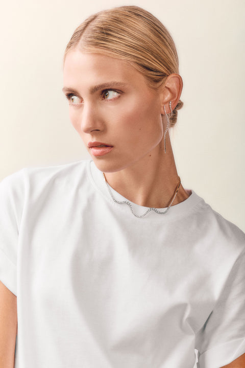 A blonde woman wearing a white crewneck t-shirt and delicate silver chain jewelry, including a scalloped necklace and long dangle earrings, set against a plain light beige background.