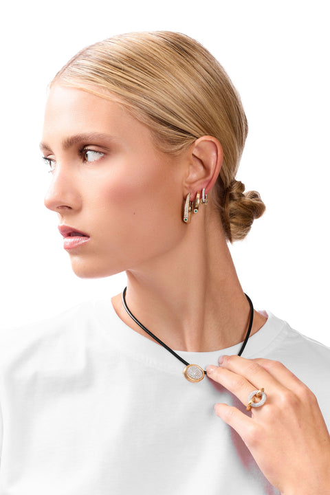 A blonde woman in profile wearing a black cord necklace with a round gold pendant featuring a clear stone, along with gold hoop earrings and a gold ring, against a solid white background.