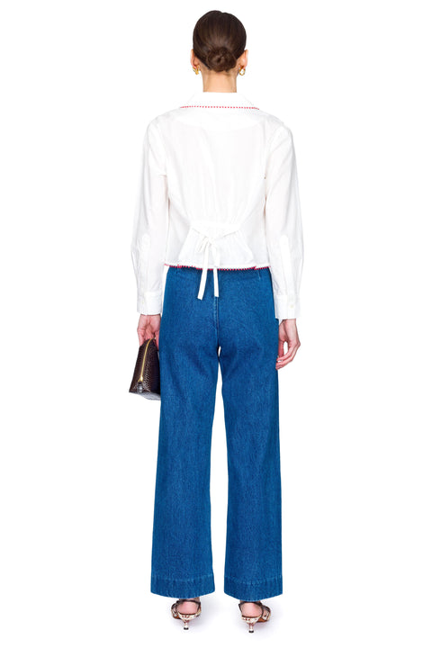 Woman wearing a white blouse with red accents and blue jeans with red buttons on a white background from the back.