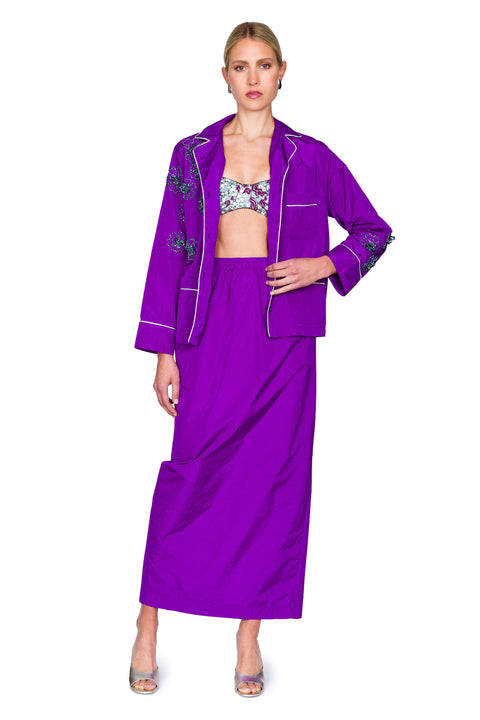 Woman wearing a purple button down pajama shirt with floral embellishments and a matching maxi skirt with one hand on her hip.