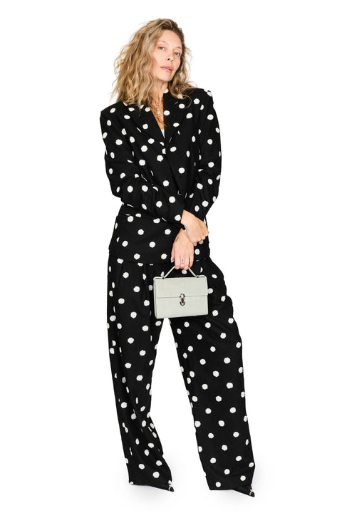 A woman modeling a black long-sleeve jumpsuit with a white polka dot pattern, holding a small light gray handbag against a plain white background.