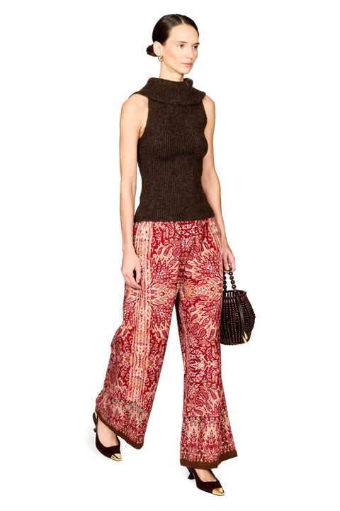 A model wearing a sleeveless dark brown knit mock-neck top and wide-leg trousers with a red and beige patterned print, holding a small black handbag against a plain white background.