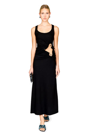 A model stands against a white background wearing a sleeveless black top and maxi skirt with a draped waist detail and a gold-toned ring accent. She is holding a small black patterned clutch and wearing blue patterned flat sandals.