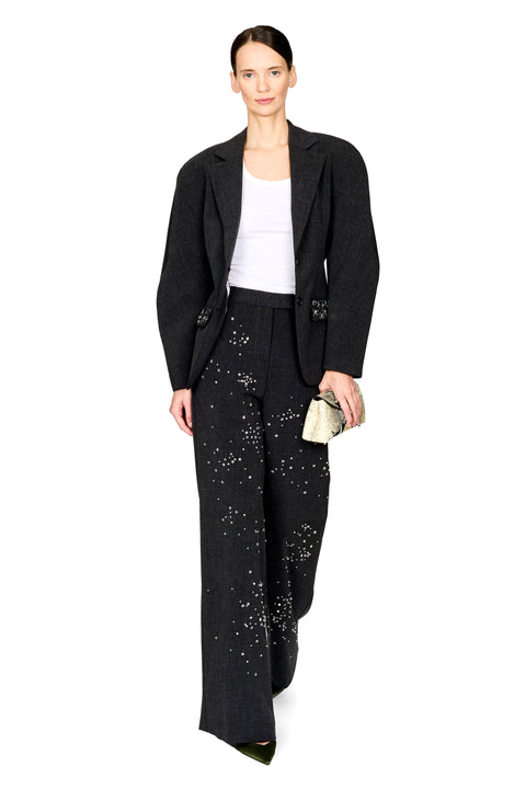 A model stands against a white studio background wearing a black textured blazer, matching wide-leg pants with silver bead embellishments, a white ribbed tank top, and holding a small metallic clutch.