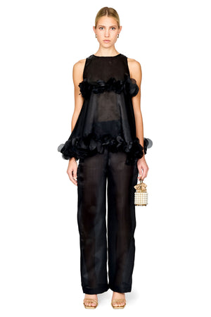 A model stands against a white studio background wearing a black sheer sleeveless top with ruffled trim and matching wide-leg sheer pants, holding a small gold-and-white woven handbag.