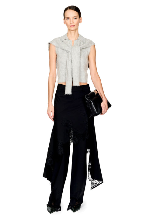 A model stands against a white studio background wearing a light gray sleeveless fuzzy wrap top with a tie detail, black wide-leg trousers with lace trim, and holding a small black clutch.