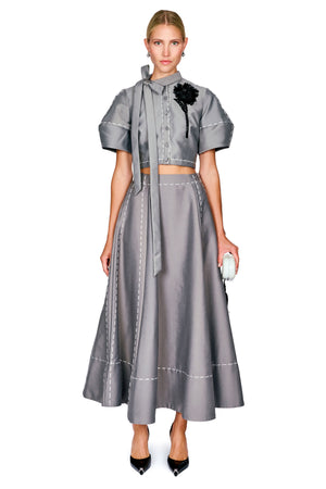 A model wearing a gray two-piece outfit consisting of a short-sleeve cropped top with a high neck and bow detail, and a matching high-waisted flared skirt with contrast stitching. She is wearing black pointed-toe heels and holding a small white clutch against a plain white background.