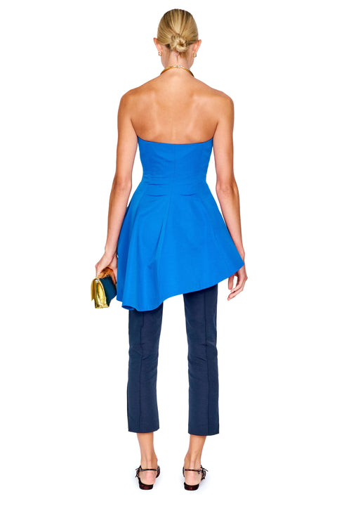 A woman shown from the back wearing a bright blue strapless peplum top, navy cropped pants, and black sandals, holding a small gold clutch against a plain white background.