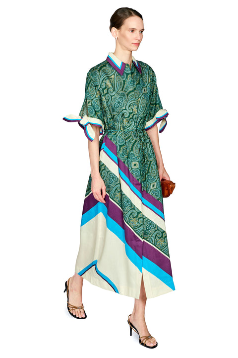 A model wearing a green patterned midi dress with purple, blue, and cream diagonal stripes, styled with brown heels and holding a small brown clutch against a white background.