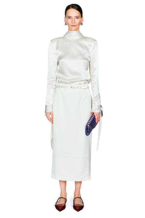 A model stands against a white studio background wearing a matching cream satin long-sleeve mock-neck top and skirt set with a matching belt, holding a purple striped clutch.