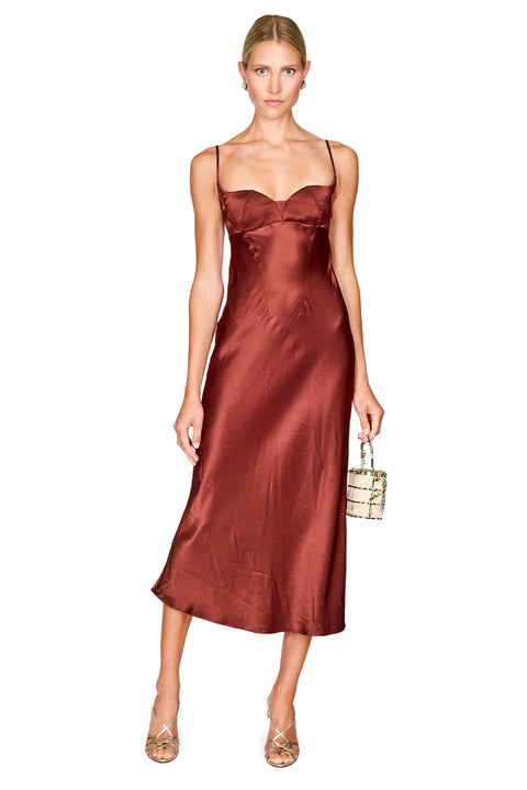 A woman modeling a satin slip midi dress in a deep rust brown color, holding a small golden handbag against a plain white background.