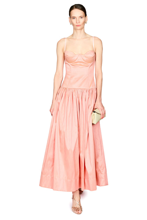 A model wearing a peach-colored sleeveless maxi dress with a structured bodice and a full, flowing skirt, holding a small beige clutch against a plain white background.