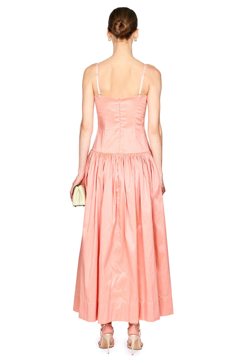 A woman shown from the back wearing a peach-pink sleeveless maxi dress with thin straps, a fitted corset-style bodice, and a gathered skirt. She is standing against a plain white background and holding a small metallic clutch.