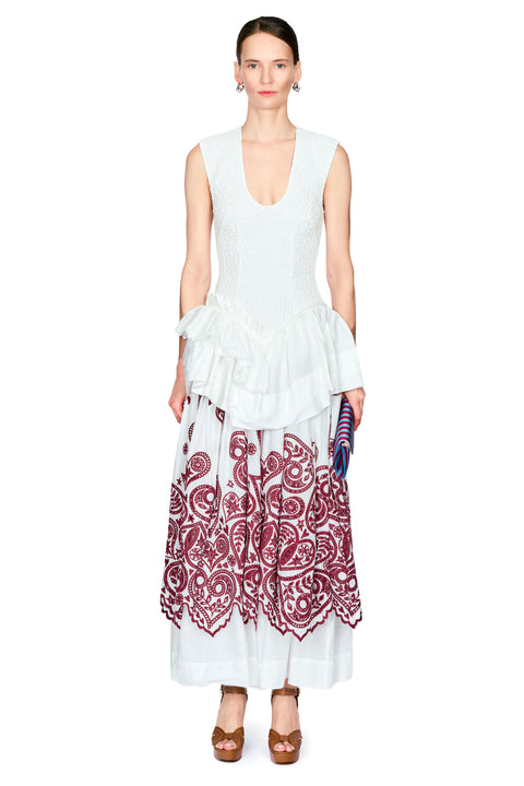 A model stands against a white studio background wearing a sleeveless white top with a ruffled waist and a long white skirt featuring a large burgundy paisley pattern.