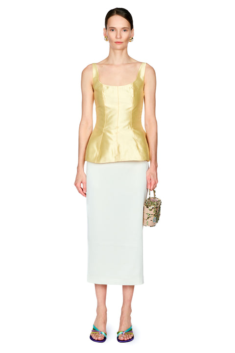 A model stands against a white studio background wearing a shiny pale yellow sleeveless top with a structured corset-style bodice and a white midi skirt, holding a small patterned handbag.